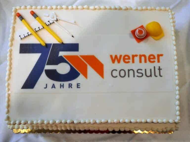 Zweistöckige, weiße Torte mit 75 Jahre WERNER CONSULT Logo bedruckt. einem Baustellenhütchen und Sicherheitshelm rechts oben. Links oben sind ein Zirkel, Bleistifte und ein Lineal.